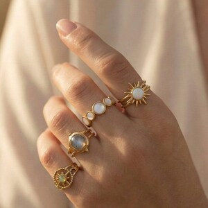 Könnte beinhalten: Ein Set aus vier goldfarbenen Ringen an den Fingern. Ein Ring hat ein Sonnenstrahlen-Design mit einem weißen Stein in der Mitte, ein anderer eine Reihe runder weißer Steine, einer zeigt einen glatten grauen Cabochon und einer einen grünen Stein.