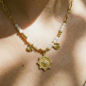 Può includere: Collana dorata con un pendente centrale con un diamante e pietre preziose circostanti. La collana ha una catena e un filo di perline bianche con dettagli dorati. Sono attaccati anche tre piccoli ciondoli rotondi dorati.