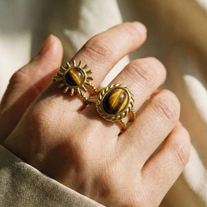 Könnte beinhalten: Zwei goldfarbene Ringe mit Tigeraugensteinen werden auf einer Hand gezeigt. Ein Ring hat ein Sonnenmuster, der andere eine seilartige Umrandung. Die Ringe sind wahrscheinlich Modeschmuck.