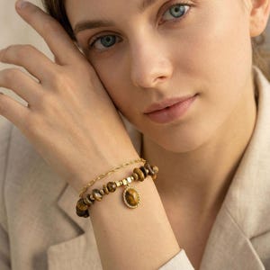 Könnte beinhalten: Zwei Armbänder sind abgebildet. Eines ist ein Perlenarmband mit braunen Tigeraugensteinen. Das andere ist ein goldenes Kettenarmband. Ein goldener Anhänger mit einem Tigeraugenstein hängt an dem Perlenarmband. Die Armbänder befinden sich am Handgelenk einer Person.