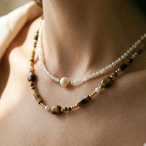 Può includere: Un set di collane a strati composto da un filo di piccole perle bianche con una perla centrale singola e un secondo filo composto da perle di occhio di tigre marrone, piccole perle bianche e distanziatori in metallo dorato.