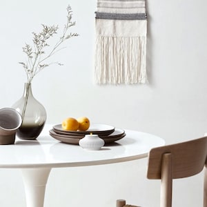 May include: A modern dining table setting with a white tabletop. Includes a gray vase with a plant, brown plates, and a small white candle holder. Two apricots sit on the plates. A woven wall hanging and a wooden chair are in the background.