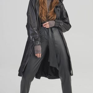 Op de afbeelding: Een zwarte jas met capuchon en een lange lijn, gecombineerd met een bijpassende zwarte broek. De jas heeft een ritssluiting en de broek heeft een uniek wikkeldetail. De outfit wordt gecompleteerd met grijs-zwarte sneakers.