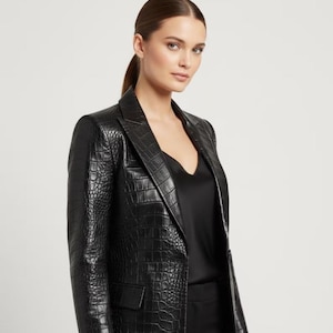 May include: A black blazer with a textured, crocodile-like pattern. The blazer has a notched lapel, a single-button closure, and a tailored fit. The model is wearing a black top underneath.