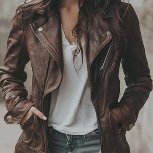 Pode incluir: Casaco de couro castanho com zíper assimétrico. O casaco tem gola entalhada, bolsos e detalhes prateados. A pessoa está vestindo uma camisa branca e jeans azul.