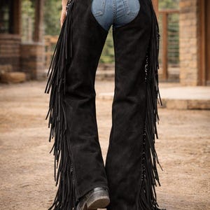 May include: Black suede chaps with long fringe detailing. The chaps are worn over blue jeans and a black top. A decorative belt with silver accents is visible. The chaps are a western-style garment.