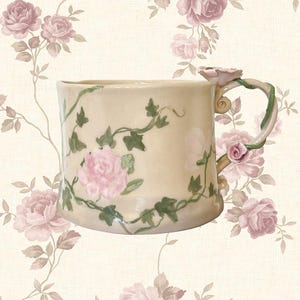 Può includere: Tazza in ceramica color crema con rose rosa e tralci verdi. Il manico è a forma di tralcio con un dettaglio di rosa rosa. La tazza è decorata con rose rosa e tralci verdi.