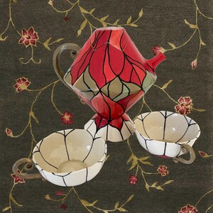 Peut inclure: Service à thé décoratif avec une théière à motifs géométriques rouges et verts et deux tasses assorties. Les tasses et la théière ont une base blanche avec des contours noirs. L'ensemble est sur un fond vert foncé avec des accents floraux.