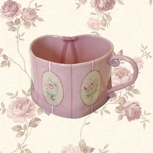 Peut inclure: Une tasse en céramique rose en forme de cœur avec une anse. La tasse présente des rayures verticales et des motifs floraux ovales. Un petit nœud orne le dessus. Le fond est un motif floral avec des roses roses.