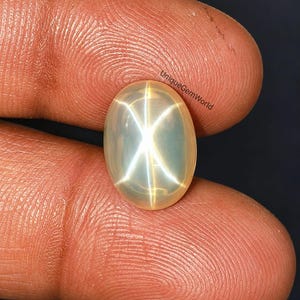 May include: An oval-shaped, translucent white star moonstone cabochon featuring a distinct six-rayed asterism effect that creates a bright, centred star pattern. The gemstone is marked with the small black text UniqueGemWorld on its surface.