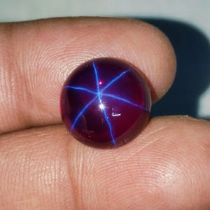 May include: A round, polished gemstone with a deep burgundy hue. The gem features a striking six-rayed star pattern in vibrant blue, radiating from the center. The stone's surface is smooth and reflective, showcasing the star effect.
