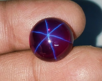 Peut inclure: Une pierre précieuse ronde et polie, d'un profond rouge bordeaux. La gemme présente un motif d'étoile à six rayons saisissant en bleu vif, rayonnant du centre. La surface de la pierre est lisse et réfléchissante, mettant en valeur l'effet d'étoile.
