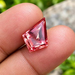 May include: A kite-shaped faceted gemstone with a vibrant pink and orange hue. The stone features precise geometric cuts that create internal light reflections and sharp edges, showcasing a transparent clarity and a polished, smooth surface finish.
