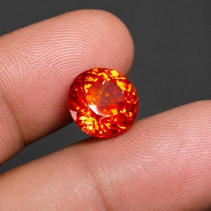 May include: A round, faceted gemstone with a vibrant orange and red color palette. The stone features a brilliant cut with numerous geometric facets that reflect light, creating a sparkling appearance against the dark, neutral background.