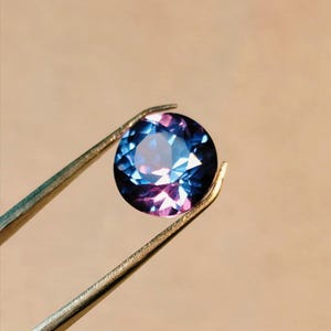 Puede incluir: Una gema redonda y facetada, sujeta con unas pinzas. La gema muestra un efecto de cambio de color, pasando del azul al púrpura. Las pinzas son de color oro metálico, sobre un fondo neutro.