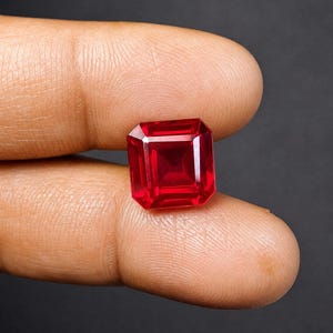 May include: A vibrant, deep red gemstone with a square, step-cut faceted design. The stone exhibits high clarity and a brilliant, reflective surface, showcasing sharp geometric edges and a symmetrical arrangement of facets that catch the light.