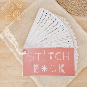 Può includere: Un libro di cucito con copertina rosa e pagine bianche a ventaglio. La copertina recita "STITCH BOOK" con illustrazioni di punti. Le pagine sono numerate e contrassegnate come "Mini stitch book". Il libro è su un panno beige.