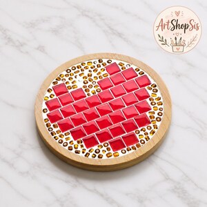 Pu&ograve; includere: Un sottobicchiere rotondo in legno con un design a mosaico a forma di cuore rosso composto da tessere di vetro quadrate. Il cuore &egrave; circondato da piccole perline color ambra su una base argentata. Un logo circolare nell'angolo riporta ArtShopSis.