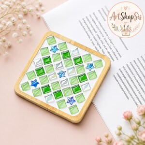 Pu&ograve; includere: Un sottobicchiere quadrato con cornice in legno chiaro caratterizzato da un motivo a mosaico di piccole tessere di vetro quadrate nei toni del verde lime, verde smeraldo e bianco iridescente, intervallate da piccole stelle di vetro blu.
