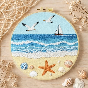 Puede incluir: Escena de playa bordada en un aro redondo. La obra presenta un cielo azul claro con dos gaviotas, un velero y olas del océano. Una playa arenosa con una estrella de mar y conchas marinas completa el diseño. El aro es de madera.