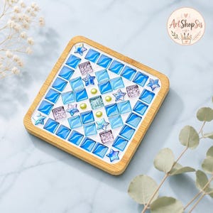 Pu&ograve; includere: Un sottobicchiere quadrato in legno con un design a mosaico composto da piastrelle quadrate blu, piastrelle a forma di stella azzurre, piccoli quadrati glitterati color lavanda e minuscole perle sferiche verde lime su base bianca.