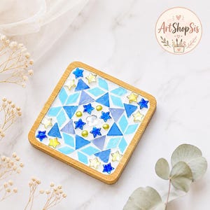 Pu&ograve; includere: Un sottobicchiere quadrato con cornice in legno caratterizzato da un design a mosaico di forme geometriche blu e azzurre, intervallate da piccoli accenti a forma di stella gialli e blu e una gemma centrale trasparente sfaccettata.