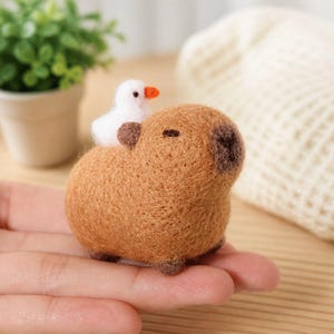 May include: A handmade felted capybara figurine in shades of brown, with a small white bird perched on its back. The bird has an orange beak. The figurine is small and detailed, showcasing a whimsical design.