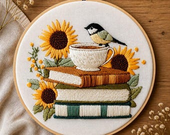May include: Embroidered hoop art featuring a stack of books, sunflowers, and a bird perched on a teacup. The embroidery uses yellow, brown, green, and white thread. The design is set against a white fabric background, framed by a wooden hoop.