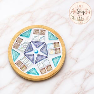 Pu&ograve; includere: Un sottobicchiere circolare in legno con un design a mosaico di tessere di vetro nelle tonalit&agrave; del viola, verde acqua e bianco iridescente. Il centro contiene una piccola perla blu circondata da pezzi di vetro geometrici a stella.