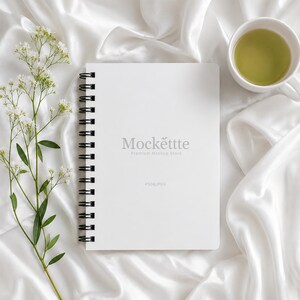 Puede incluir: Un cuaderno de espiral blanco con encuadernación de alambre negro. La portada presenta el texto gris Mockette Premium Mockup Store y PSD&JPEG. Una rama de pequeñas flores blancas y una taza de cerámica blanca con té verde claro están presentes.