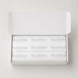 Könnte beinhalten: Ein weißer, rechteckiger Versandkarton aus Pappe mit offenem Deckel, der gefaltetes weißes Seidenpapier enthält, welches mit dem sich wiederholenden grauen Text Mockettte Premium Mockup Store in einem Rastermuster bedruckt ist.