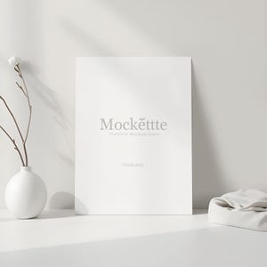 May include: A minimalist mockup scene featuring a white canvas with the text "Mockettte Premium Mockup Store" and "PSD & JPEG". A white vase with a sprig of flowers and a folded cloth are also present.