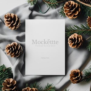 May include: A white card with the text "Mockette Premium Mockup Store" and "PSD & JPEG" is surrounded by pine cones and evergreen branches on a gray cloth. The pine cones are brown and the branches are green.