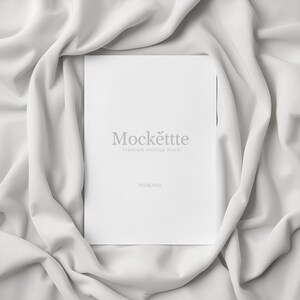 May include: A white rectangular card with the text "Mockëttte Premium Mockup Store" and "PSD&JPEG" in a soft, flowing white fabric. The card is centered, with the fabric draped around it.