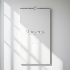 Può includere: Un mockup di calendario rettangolare bianco appeso a una parete bianca. Il calendario ha una rilegatura a spirale nella parte superiore e un gancio di metallo. Il testo "Mockëttte Premium Mockup Store" è stampato sul calendario. Il testo "PSD & JPEG" è stampato in basso.