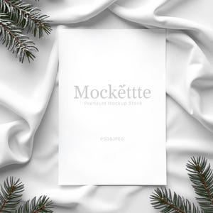 May include: A white rectangular card with the text "Mockettte Premium Mockup Store" and "PSD & JPEG" is centered on a white satin fabric. Green pine branches are in the corners. The image is a mockup for stationery or invitations.