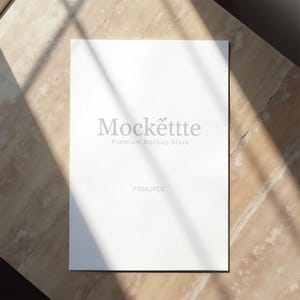 May include: A white rectangular paper with the text "Mockettte Premium Mockup Store" in gray. The paper is on a light brown surface with shadows. The text "PSD & JPEG" is at the bottom of the paper.
