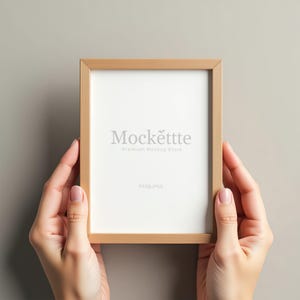 Puede incluir: Un marco de fotos rectangular de madera marrón claro con interior blanco, sostenido por dos manos. El marco tiene el texto "Mockette" y "Premium Mockup Store" impreso en el interior blanco. El fondo es gris neutro liso.