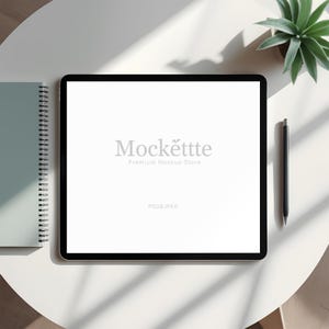 May include: A digital tablet with a black bezel, displaying the text "Mockëttte Premium Mockup Store". A black stylus, a spiral-bound notebook, and a small potted succulent are also visible on a white surface.