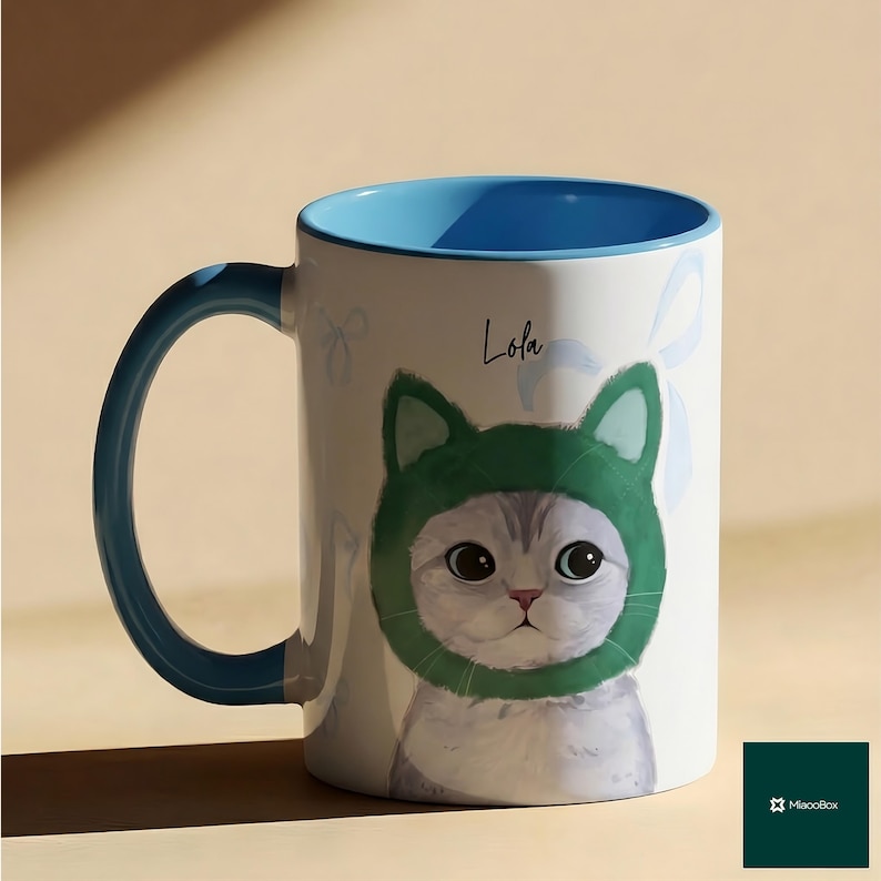 May include: White ceramic mug with a blue interior and handle. The mug features a cat wearing a green hat, with the name "Lola" written above the cat. The mug has a light blue bow design.