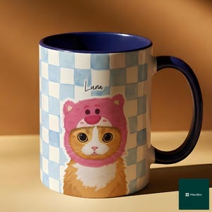 May include: A ceramic mug with a blue interior and handle, featuring a cat wearing a pink bear hat. The mug has a light blue and white checkered pattern and the name "Luna" is written on the side.