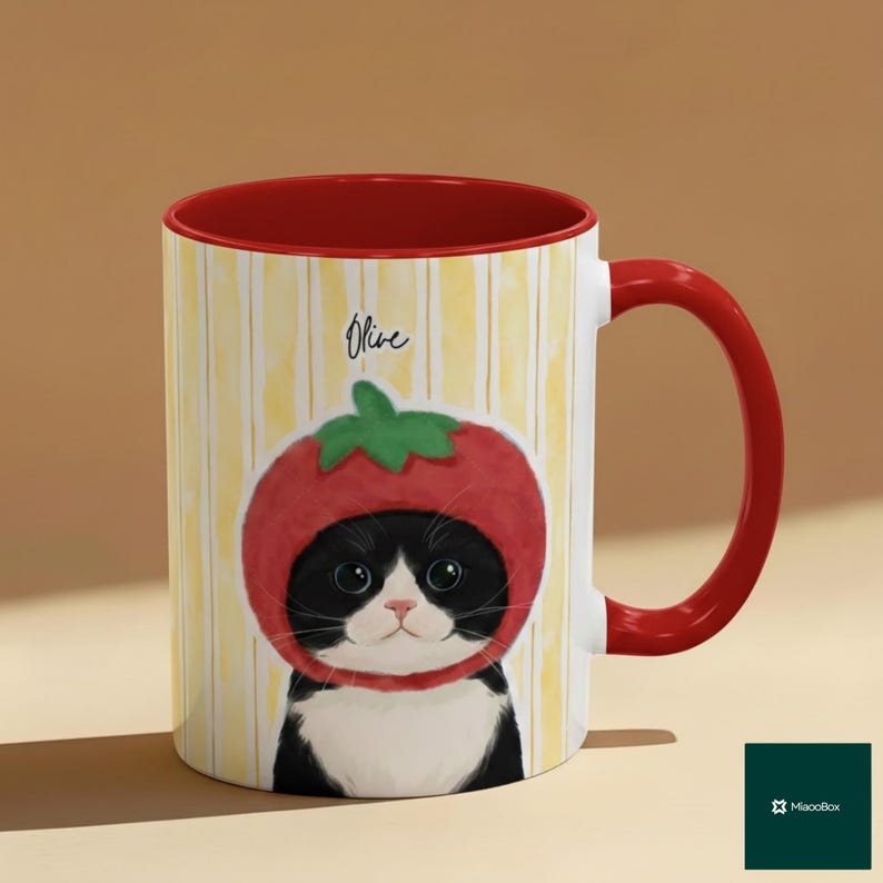 May include: A ceramic mug with a red interior and handle. The mug features a whimsical illustration of a black and white cat wearing a tomato hat. The mug has the name "Olive" written on it, and a yellow striped background.