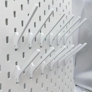 Op de afbeelding: Een wit pegboard-paneel met een raster van verticale sleuven, uitgerust met meerdere witte schuine haken die aan het oppervlak zijn bevestigd. De haken zijn gerangschikt in twee horizontale rijen en vormen een modulair systeem voor opslag.