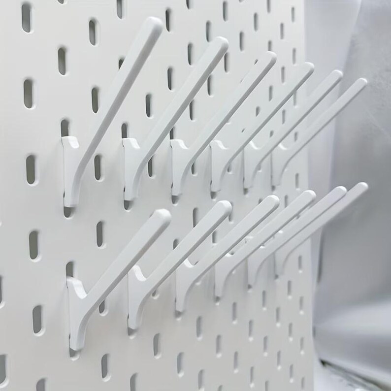 Op de afbeelding: Een wit pegboard-paneel met een raster van verticale sleuven, uitgerust met meerdere witte schuine haken die aan het oppervlak zijn bevestigd. De haken zijn gerangschikt in twee horizontale rijen en vormen een modulair systeem voor opslag.
