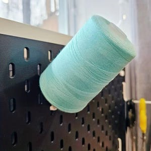 Op de afbeelding: Een grote klos licht mintgroen naaigaren gemonteerd op een zwart geperforeerd bord. De cilindrische klos heeft een glad, strak gewonden oppervlak van garen en een witte plastic kern aan de bovenkant, geplaatst tegen het donkere paneel.