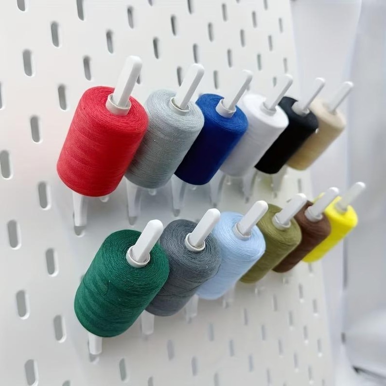 Op de afbeelding: Een witte geperforeerde wandorganizer met twaalf klossen naaigaren in diverse kleuren, waaronder rood, grijs, blauw, wit, zwart, beige, groen, donkergrijs, lichtblauw, olijfgroen, bruin en geel, elk bevestigd op een witte kunststof pin.