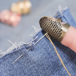 May include: A metallic bronze-toned open-sided thimble with a dimpled surface texture, worn on a finger while pushing a silver sewing needle through a piece of blue denim fabric with frayed edges and a single strand of yellow thread attached.