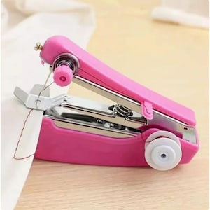 May include: A handheld portable sewing machine in a bright pink plastic casing with silver metal components. The device features a white plastic bobbin on the side and a needle mechanism currently stitching through a piece of white fabric.