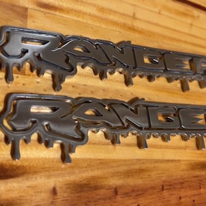 May include: Two metallic silver "RANGER" emblems with a dripping design are displayed on a wooden surface. The word "RANGER" is spelled out in a stylized font, with the dripping effect adding a unique visual element to the emblems.