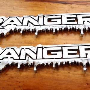 Può includere: Due emblemi "RANGER" color argento con un design a goccia. La parola "RANGER" è in maiuscolo nero in grassetto. Gli emblemi sono su una superficie di legno.